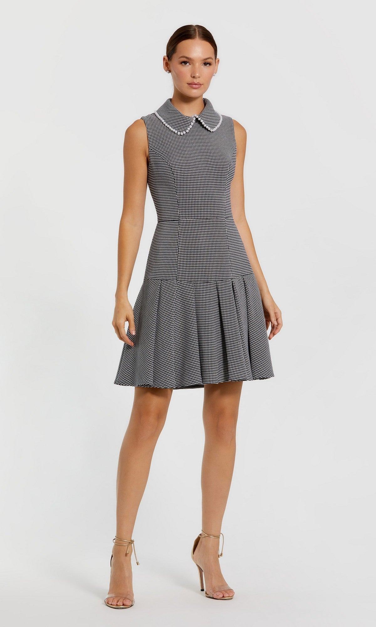 A woman stands against a plain light background wearing the Mac Duggal 27795 Short Wedding Guest Dress in black and white, featuring a sleeveless, knee-length checkered design with a pearl embellished collar, paired with beige heeled sandals.